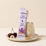A Kids Dream PM sachet torn open, honey running onto a spoon, cherries and a chamomile flower beside it